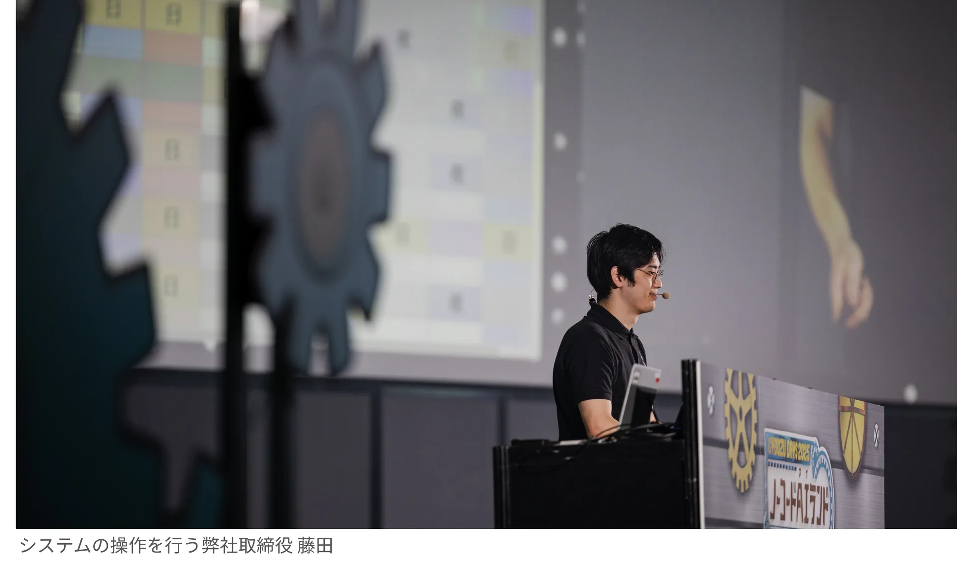 演台でシステム操作を行うフリグラム取締役 藤田。背後のスクリーンにシフト管理の画面が映し出されている。