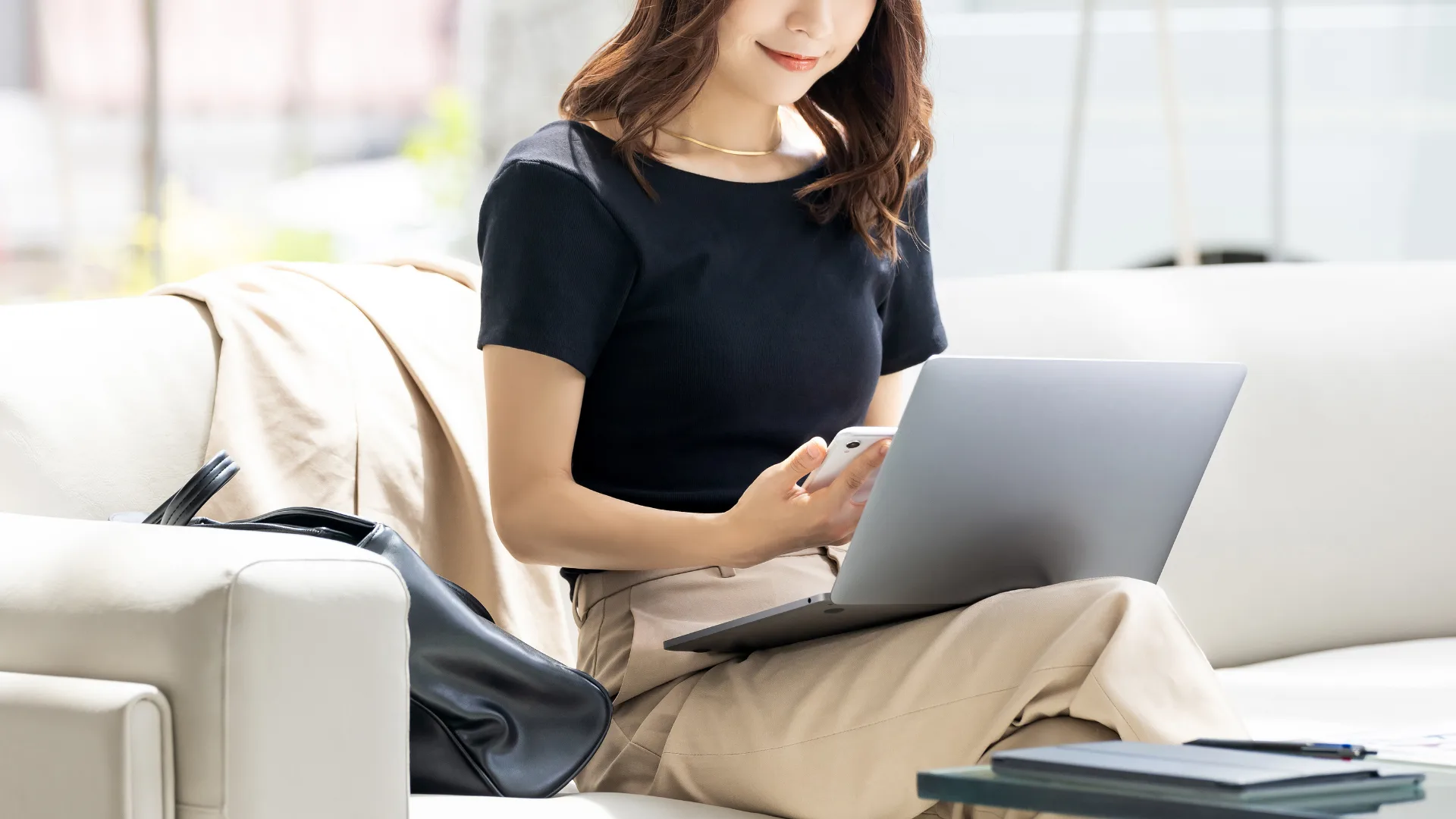 ソファでPCとスマホを操作する女性、電子化・リモートワークのイメージ