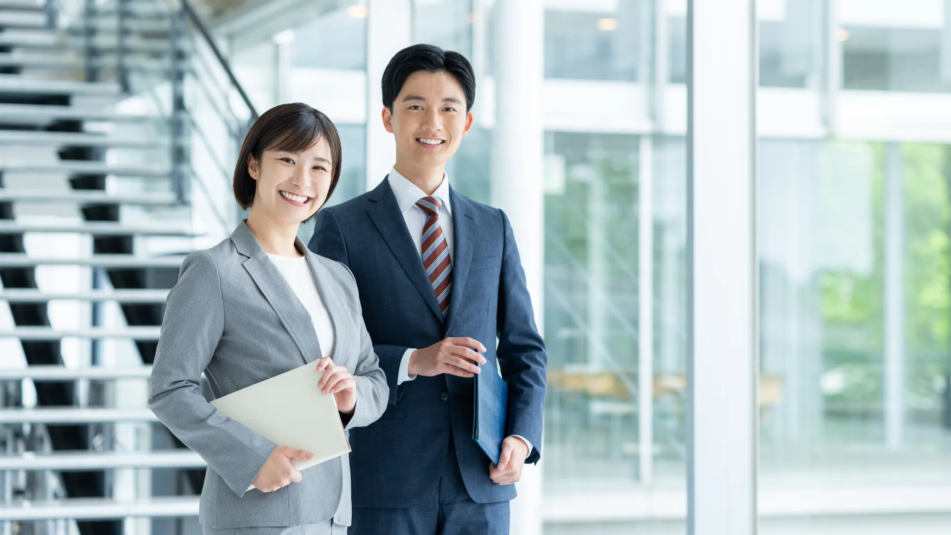オフィスの窓際で笑顔で立つ男女のビジネスパーソン。