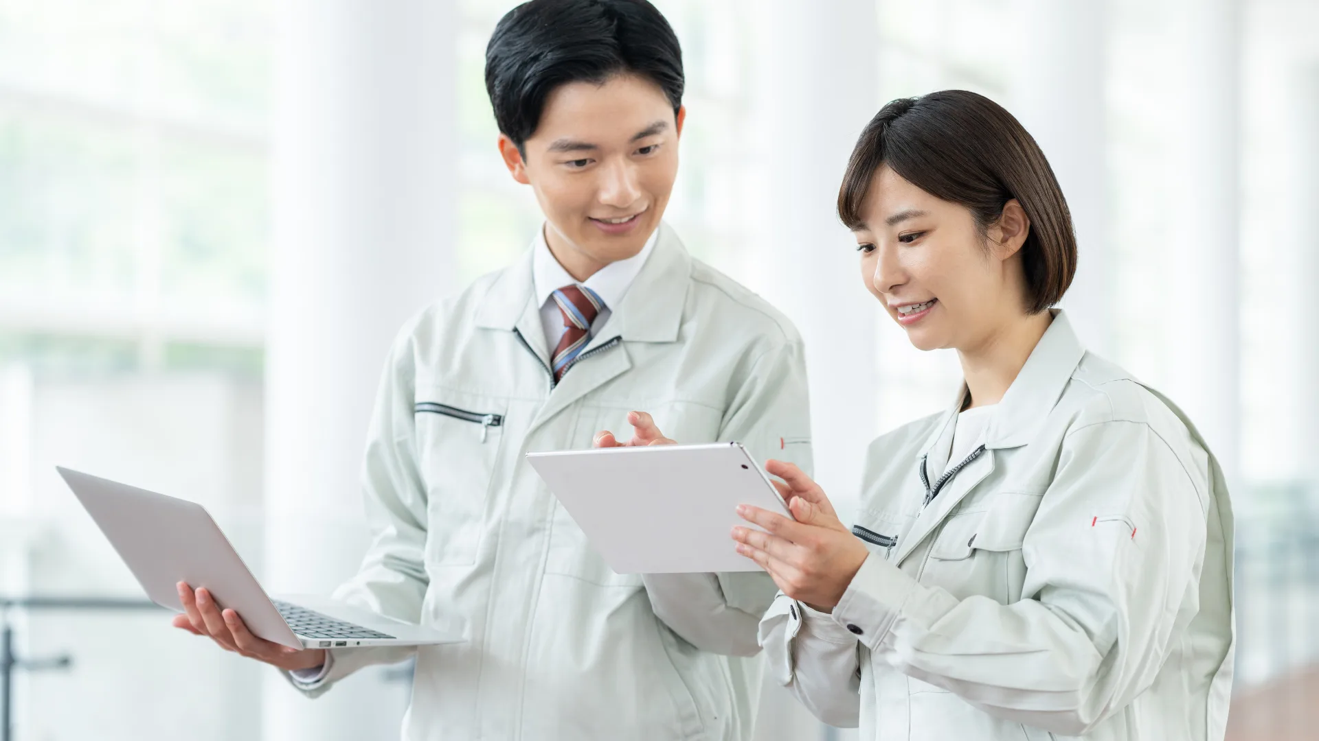 作業着姿の男女二人がパソコンとタブレットで業務改善に向けて相談している。