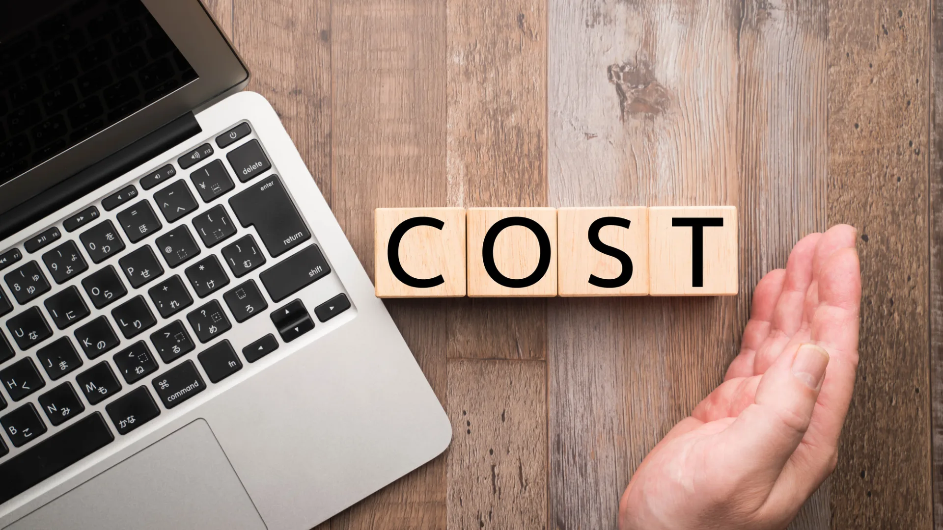 ノートパソコンの横に「COST」と書かれたブロックがあり、手が添えられている画像。