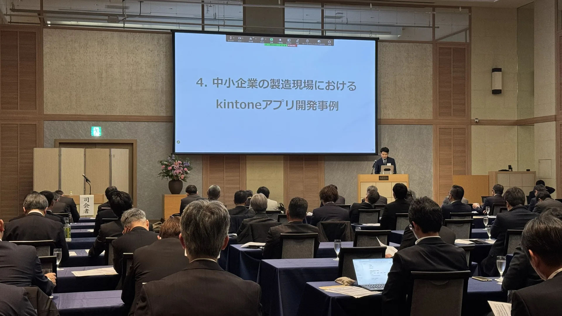 登壇中の様子。スクリーンに「中小企業の製造現場におけるkintoneアプリ開発事例」のスライドが映し出され、多くの参加者が聴講している。