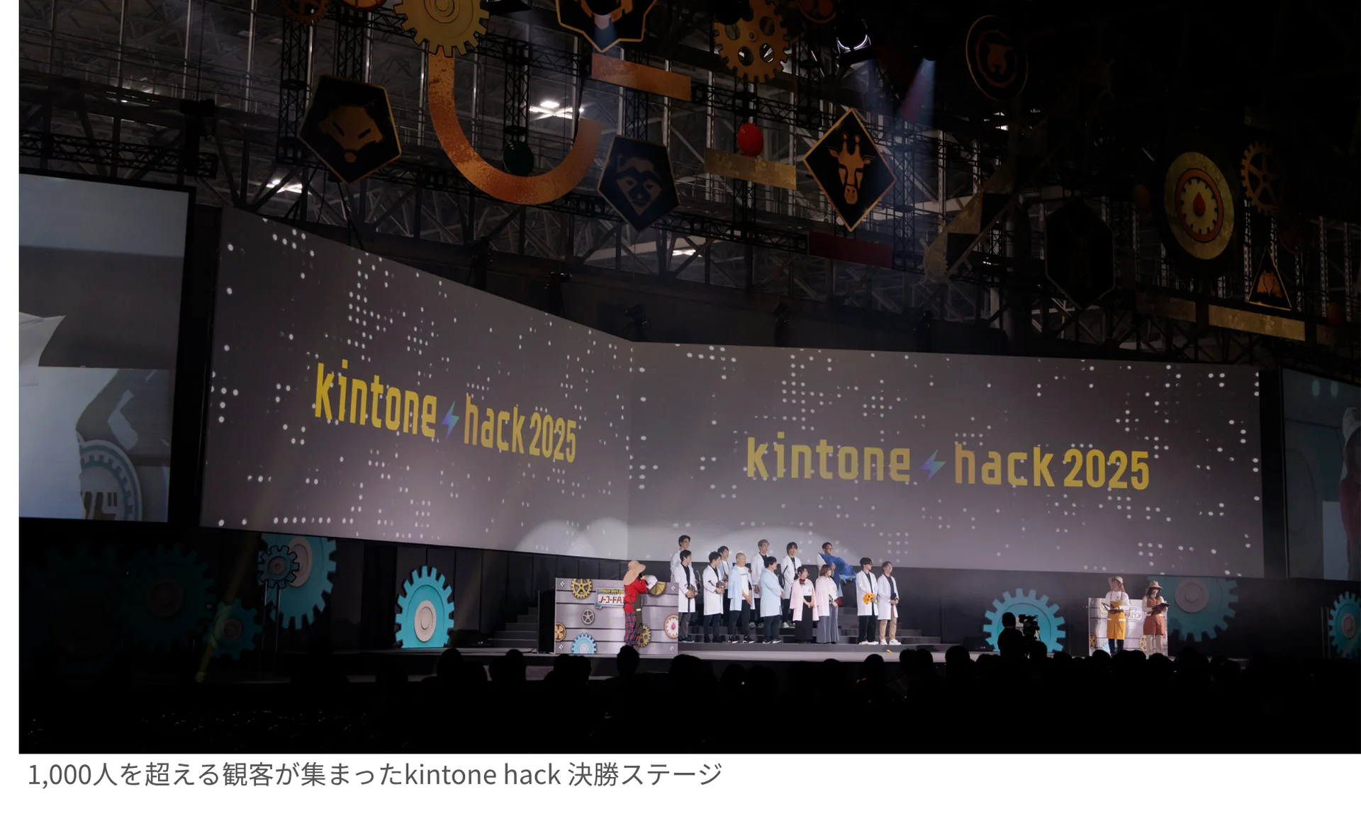 kintone hack 2025の決勝ステージ全景。大型スクリーンにイベントロゴが映し出され、ファイナリストたちがステージに並んでいる。
