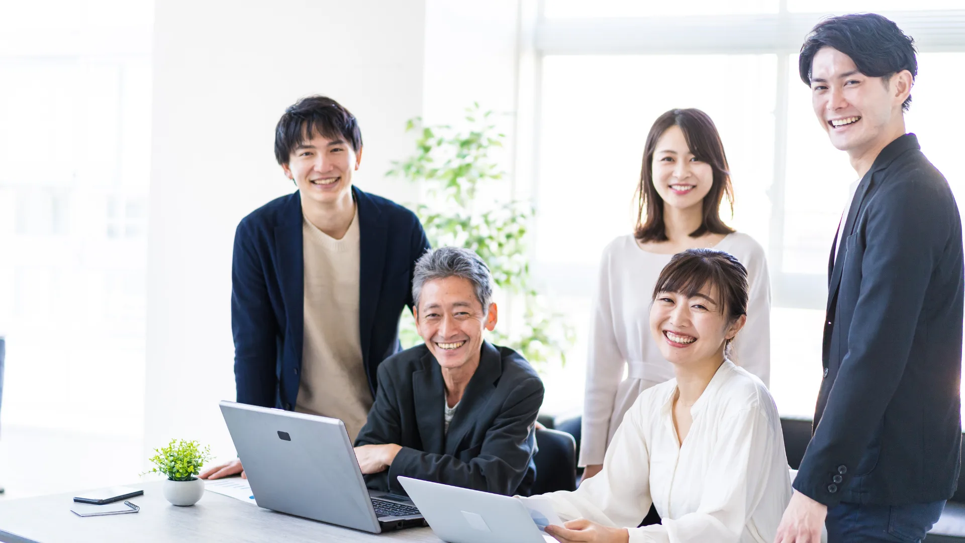 オフィスでPCを囲み、笑顔でカメラを見るビジネスチーム