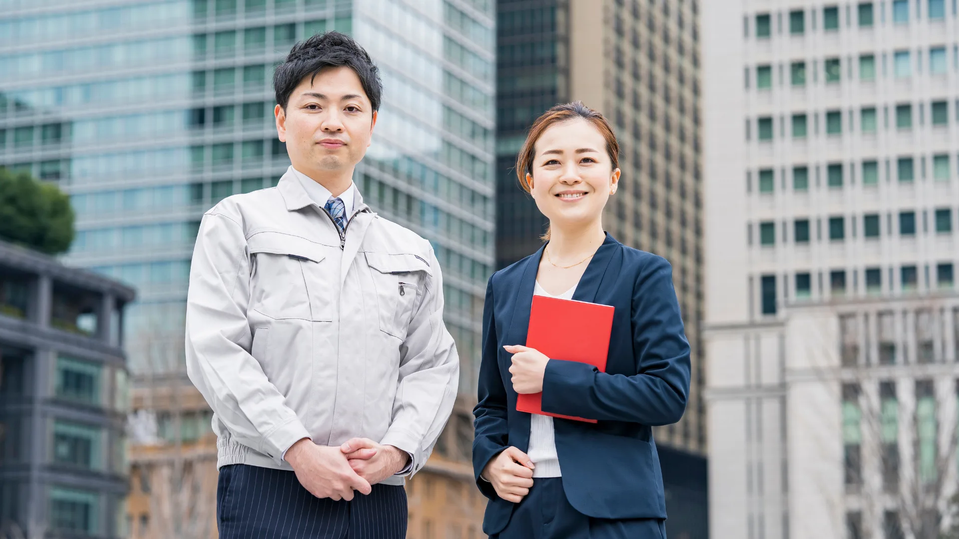 ビル群を背景に、作業服とスーツの男女が立つビジネスパーソン